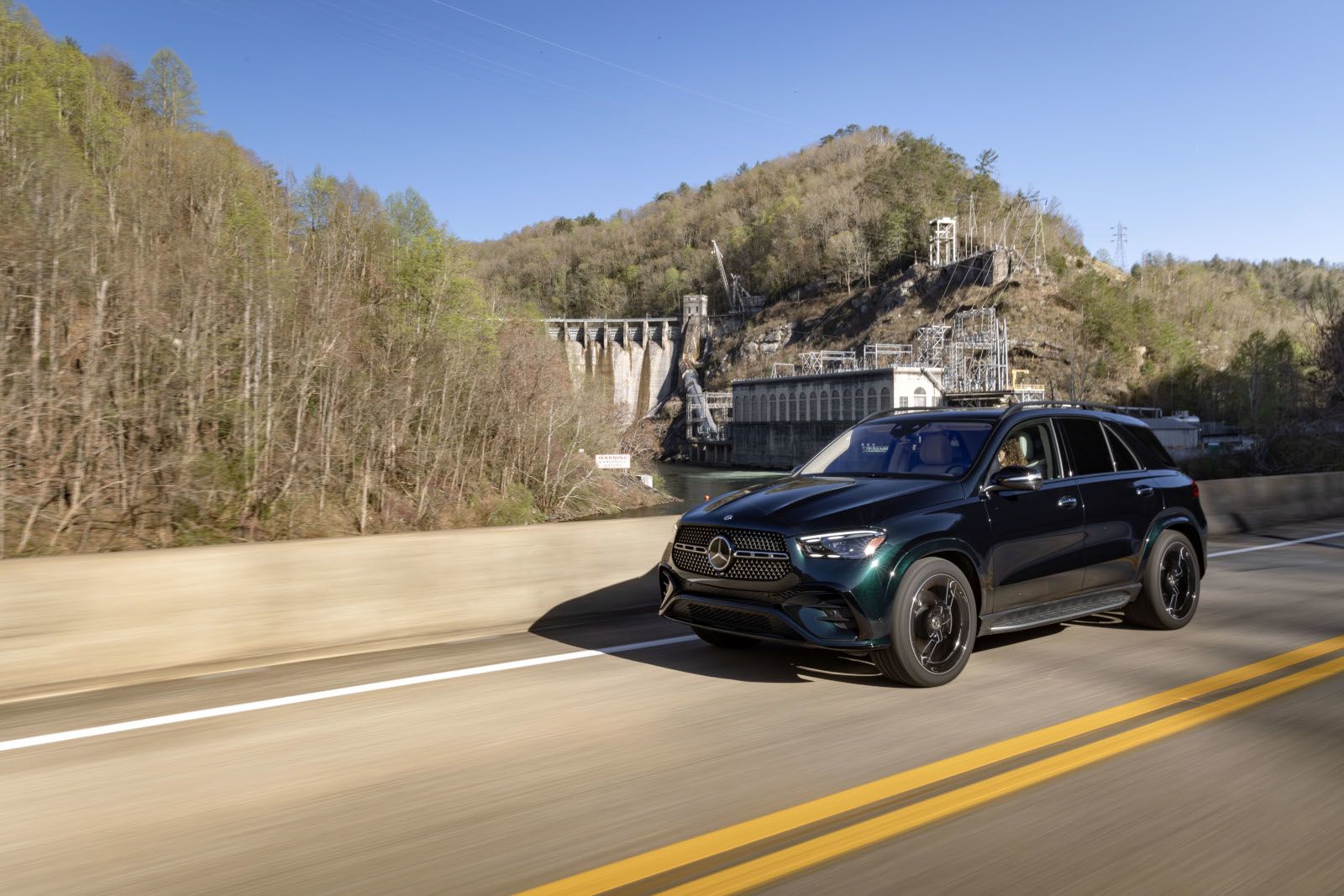 2026 Mercedes-Benz GLE 450 - A dark green 2026 Mercedes-Benz GLE 450 driving on a highway alongside a river and a dam - Mercedes-Benz Brampton - New car dealership in Brampton, Ontario