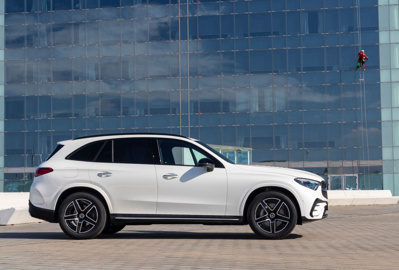 2026 Mercedes-Benz GLC - Side profile view of a white 2026 Mercedes-Benz GLC SUV parked in front of a modern glass skyscraper - Mercedes-Benz Brampton - New car dealership in Brampton, Ontario