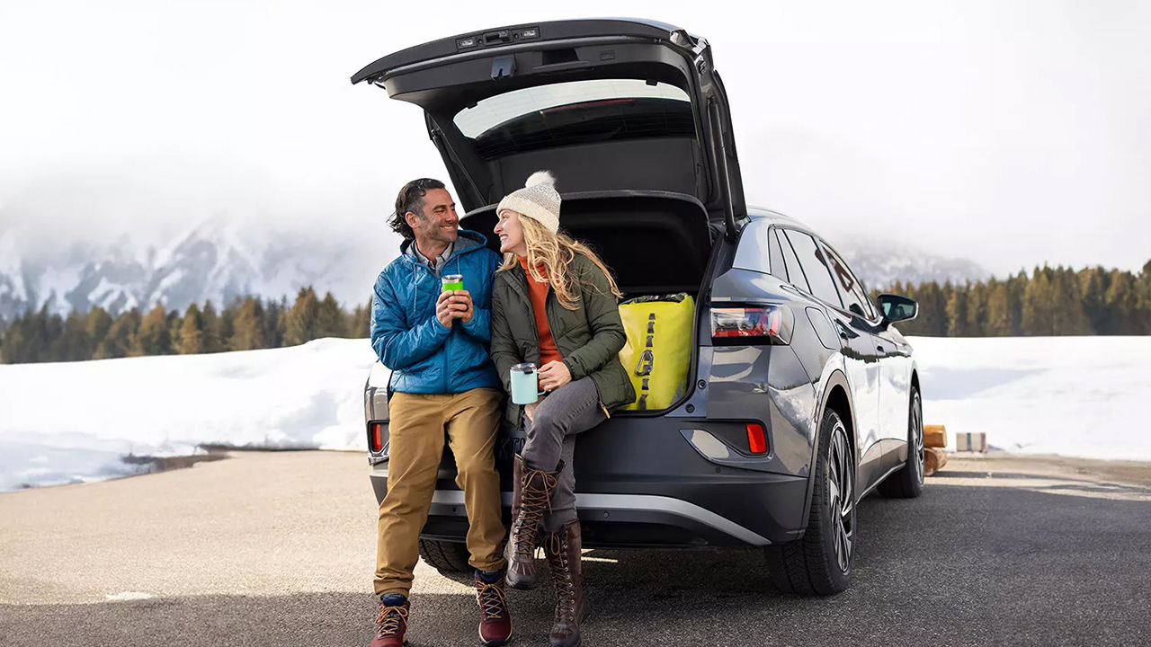 Vue arrière du Volkswagen ID.4 2024 en hiver stationné avec valise ouverte et un couple prenant un café chaud