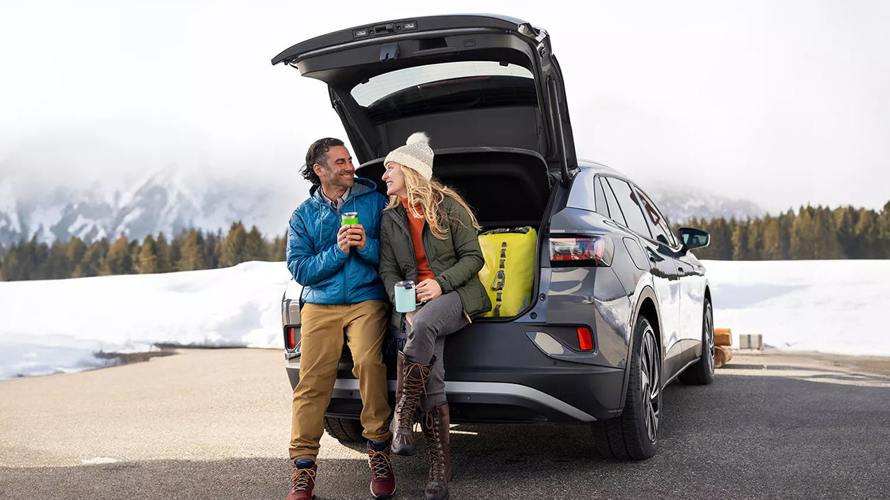 Rear view of Volkswagen ID.4 2024 parked in winter with suitcase open and a couple enjoying a hot cup of coffee.