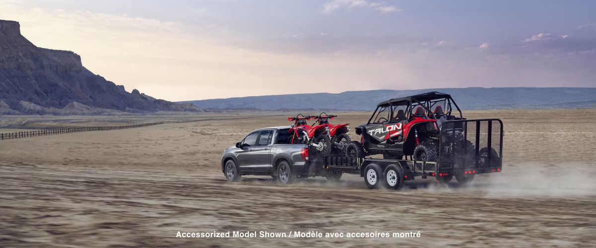 vue latérale arrière d'une Honda Ridgeline 2025 dans le désert avec deux motos tout terrain dans la caisse et qui traine une remorque avec un VTT