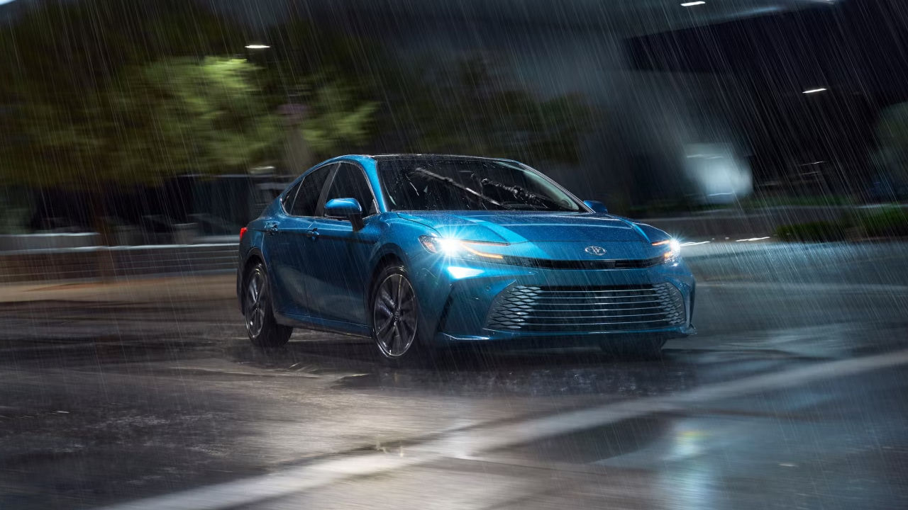 3/4 front and side view of the Toyota Camry hybrid 2025, parked with headlights on on a rainy day.