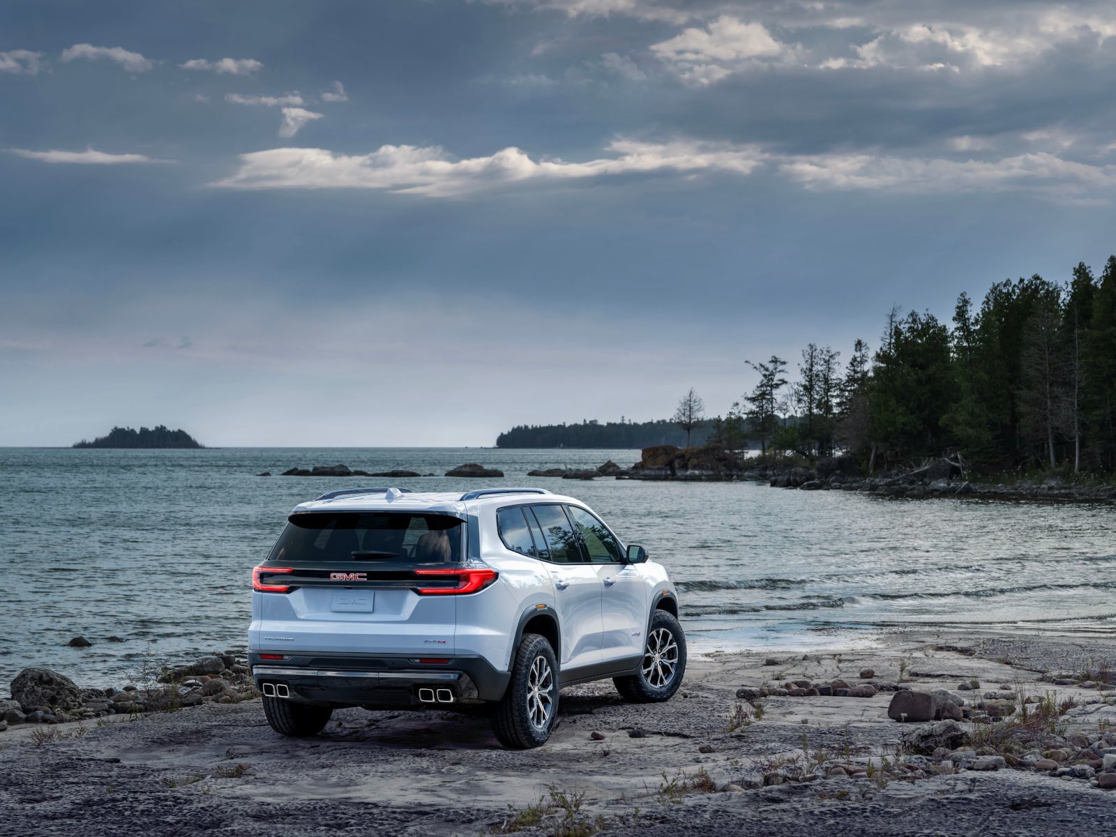 2025 GMC Acadia AT4 - rear view of a white 2025 GMC Acadia AT4 parked on a rocky shore next to the water - Applewood Chevrolet Buick GMC - GMC car dealership in Mississauga, Ontario