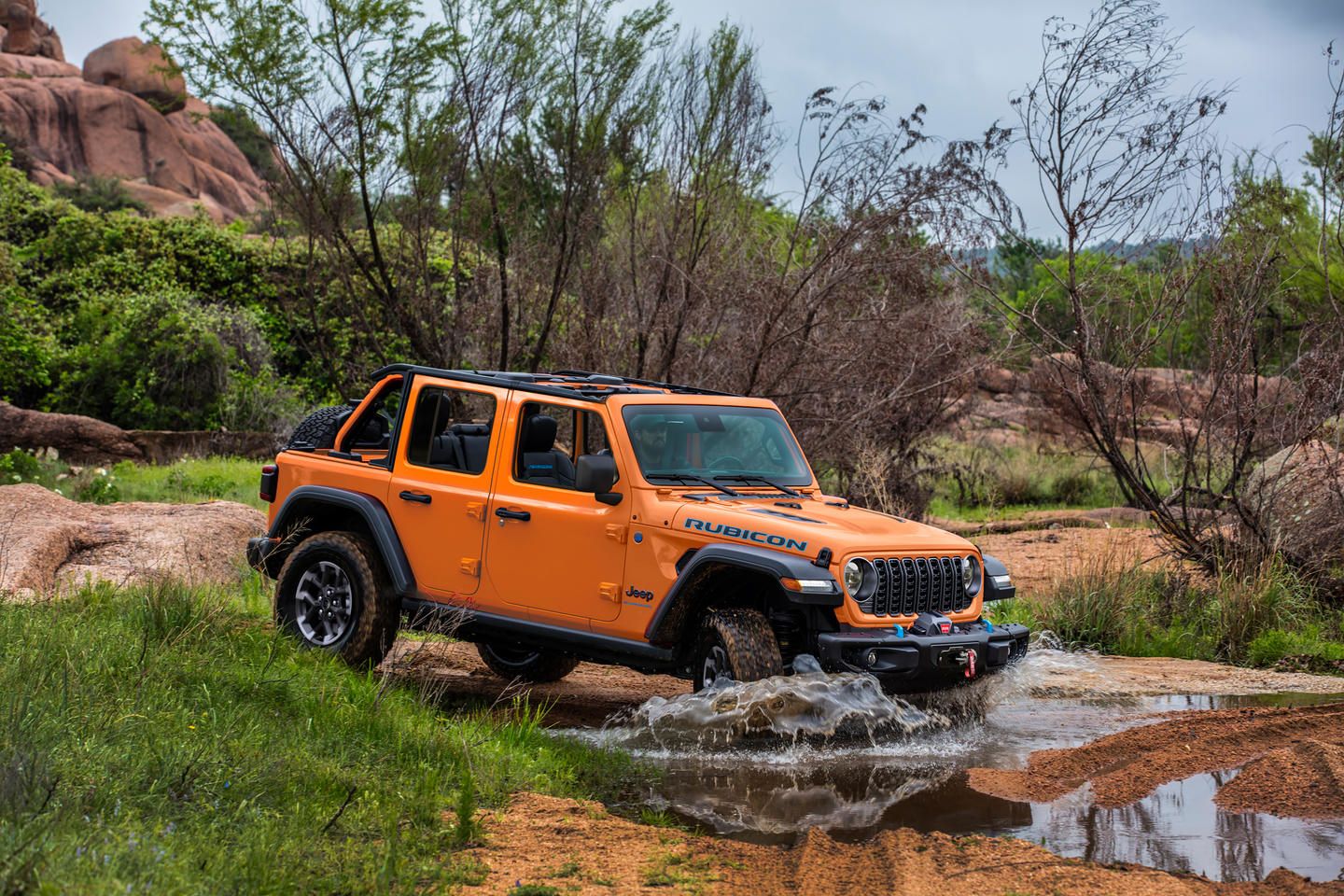 2025 Jeep Wrangler Rubicon - an orange 2025 Jeep Wrangler Rubicon driving through a stream in a grassy, wooded area - Cooksville Dodge Chrysler Jeep Ram - Jeep car dealership in Mississauga, Ontario