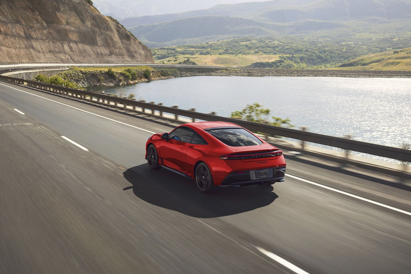 2026 Honda Prelude - Rear three-quarter view of a red Prelude driving on a scenic highway. - Orangeville Honda - New car dealership in Orangeville, Ontario