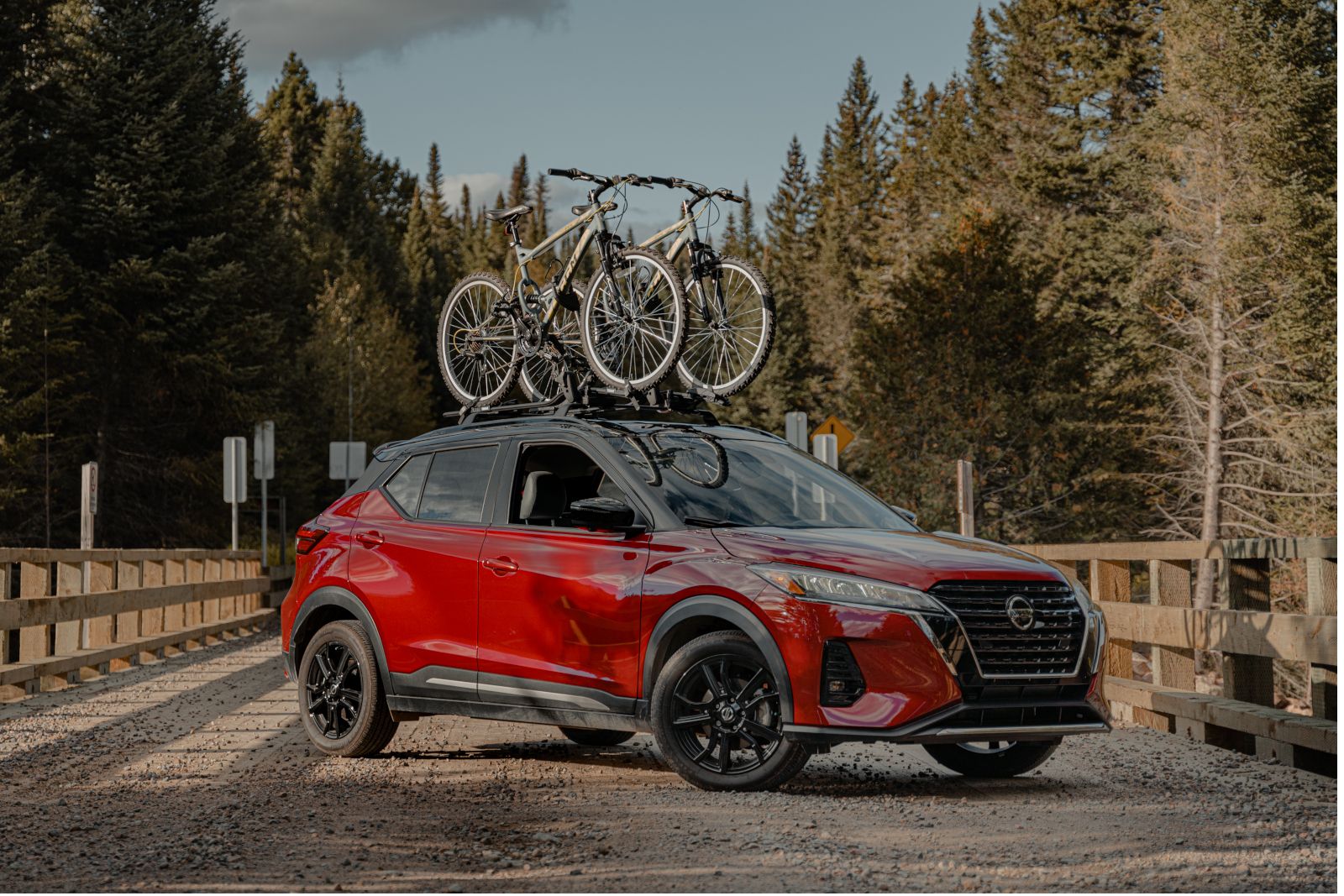 Nissan Kicks - A red Nissan Kicks with a black roof and a bike rack, parked on a gravel bridge surrounded by trees - AutoPark Barrie - Used car dealership in Barrie, Ontario