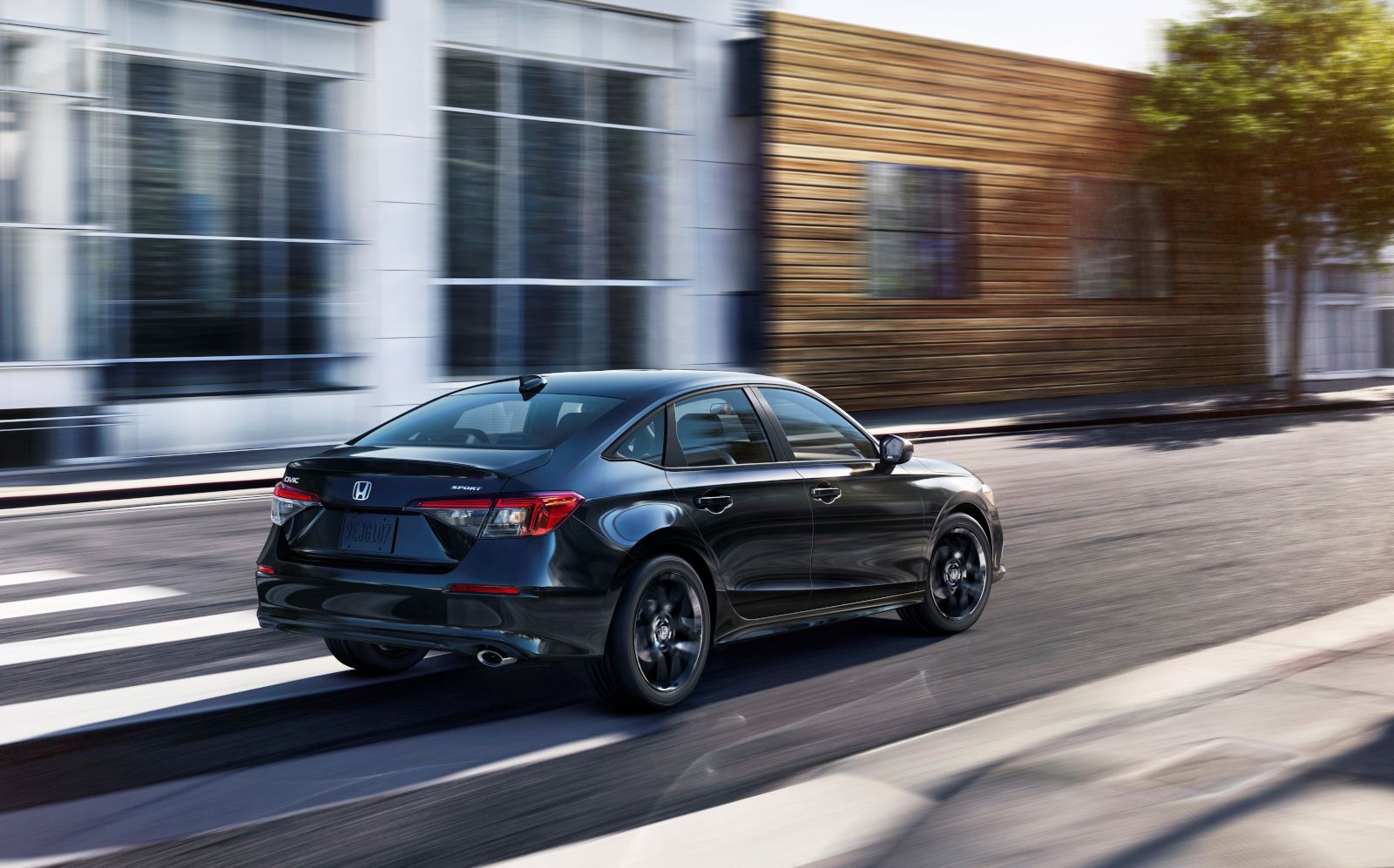2026 Honda Civic Sedan - Rear three-quarter view of a black Honda Civic Sedan driving down a city street - Orangeville Honda - Honda car dealership in Orangeville, Ontario