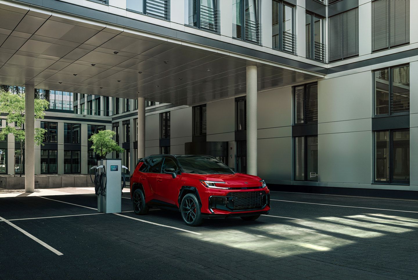 2026 Toyota RAV4 PHEV - a red 2026 Toyota RAV4 PHEV plugged into a charging station outside a modern building - Oakville Toyota - New car dealership in Oakville, Ontario