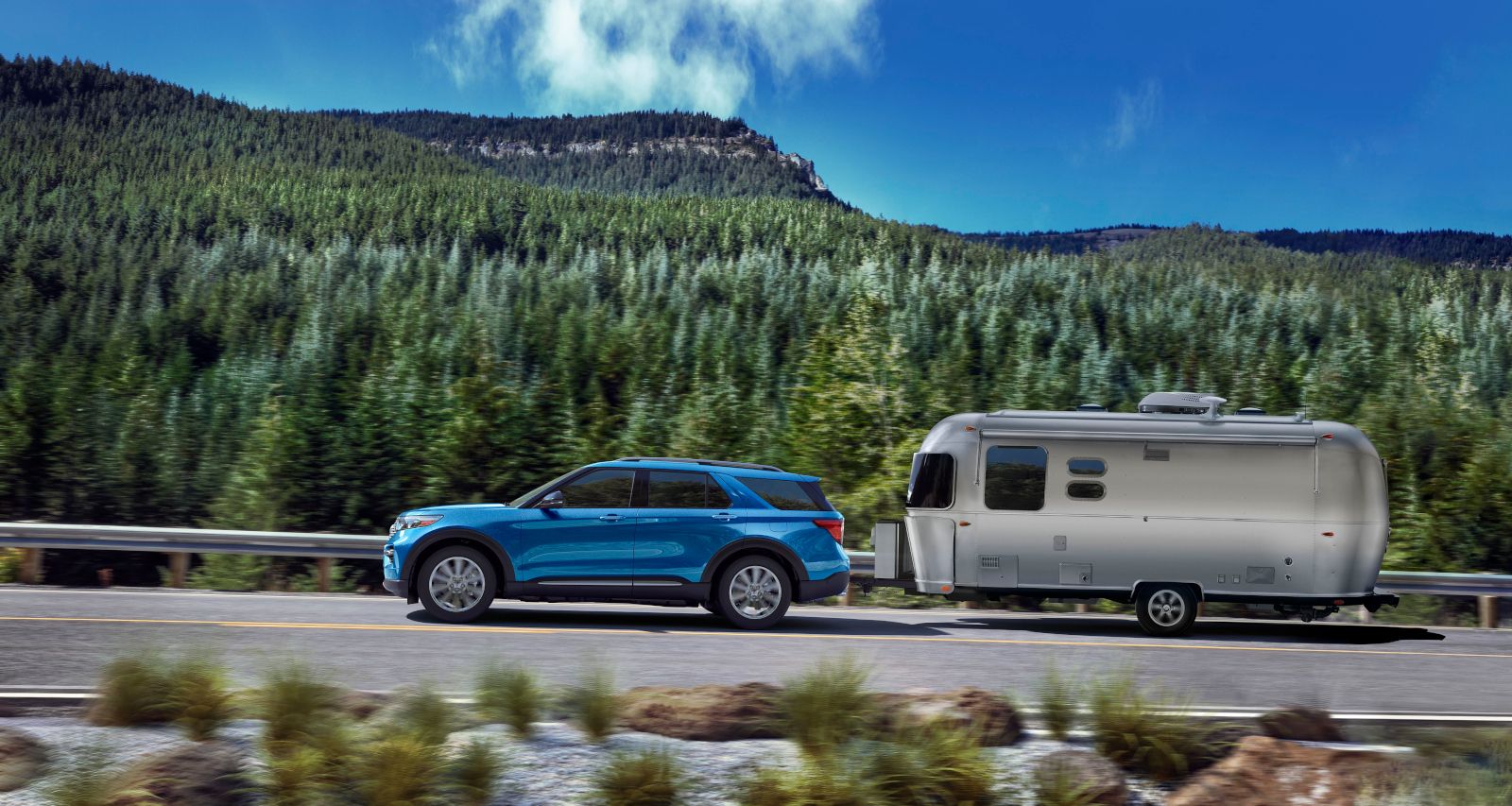 Ford Explorer Hybrid - a blue Ford Explorer Hybrid towing a silver camping trailer on a scenic highway - Autopark Barrie - Used car dealership in Barrie, Ontario