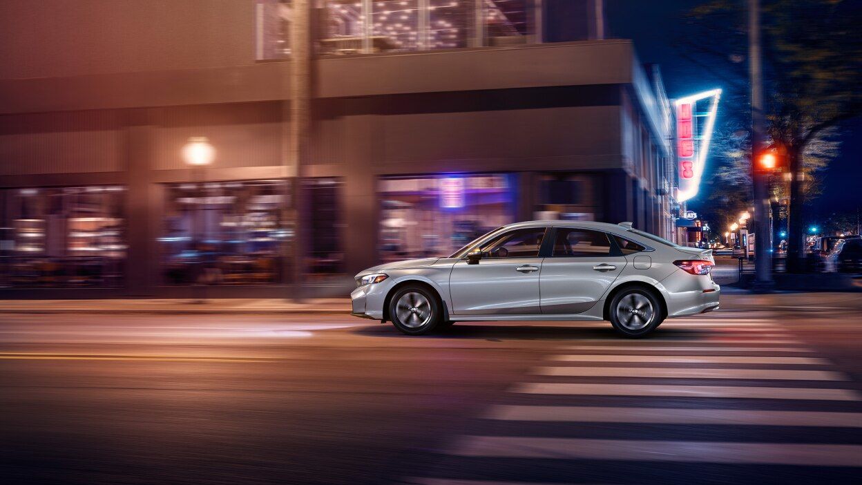 2026 Honda Civic Sedan &ndash; a silver Honda Civic LX Sedan driving through a city street at night with motion blur and illuminated storefronts - Orangeville Honda - New car dealership in Orangeville, Ontario