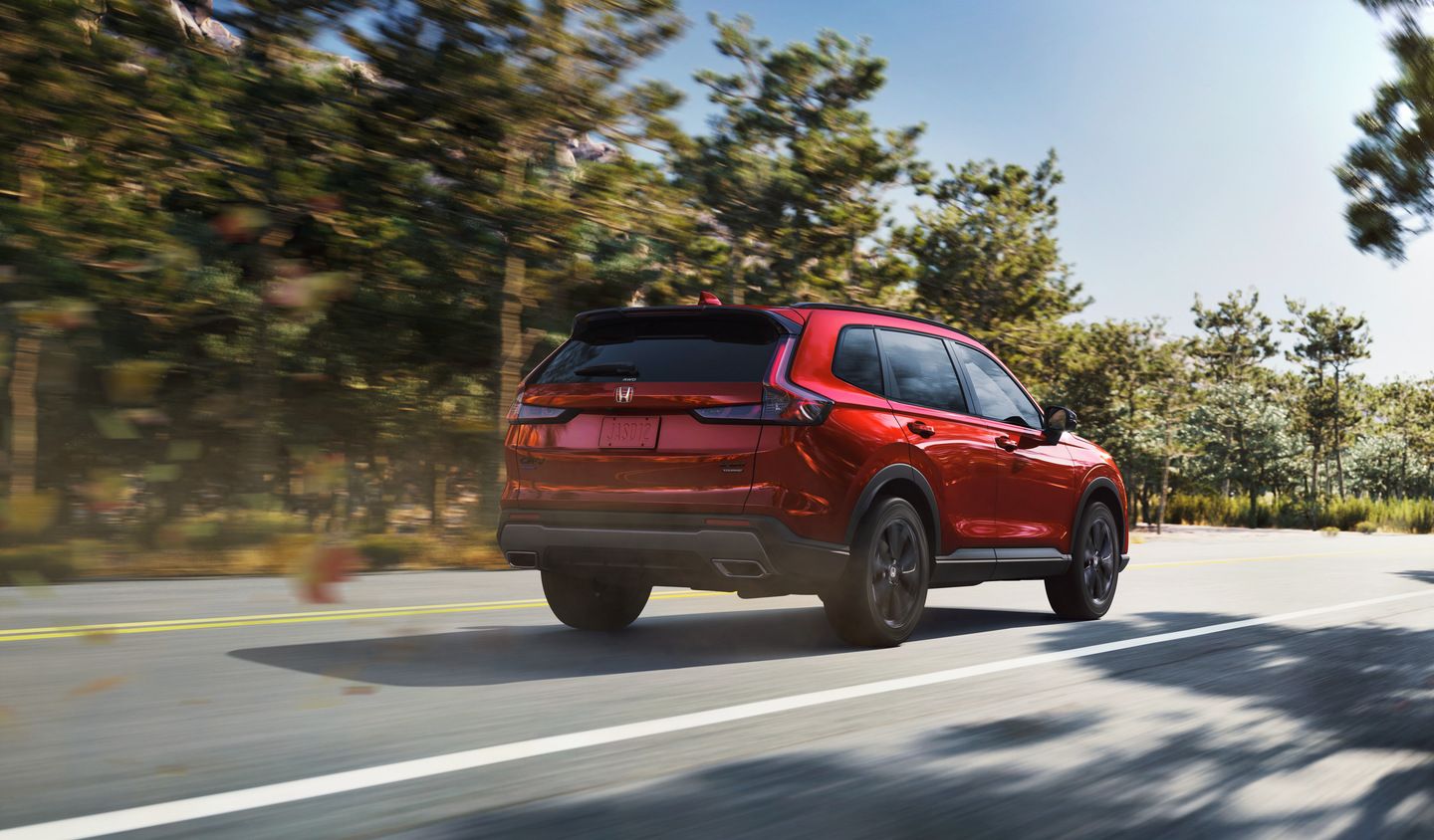 2026 Honda CR-V Sport Touring Hybrid - rear 3/4 view of a red Honda CR-V Sport Touring Hybrid driving on a paved road - Orangeville Honda - New car dealership in Orangeville, Ontario