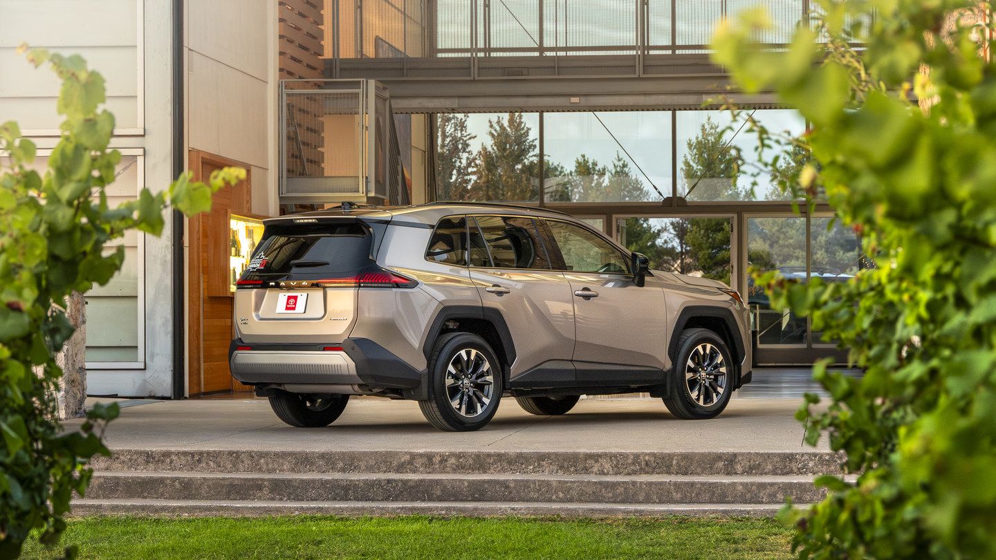 2026 Toyota RAV4 - Rear three-quarter view of a bronze Toyota RAV4 parked outside a modern glass building framed by green foliage - Georgetown Toyota - New car dealership in Georgetown, Ontario