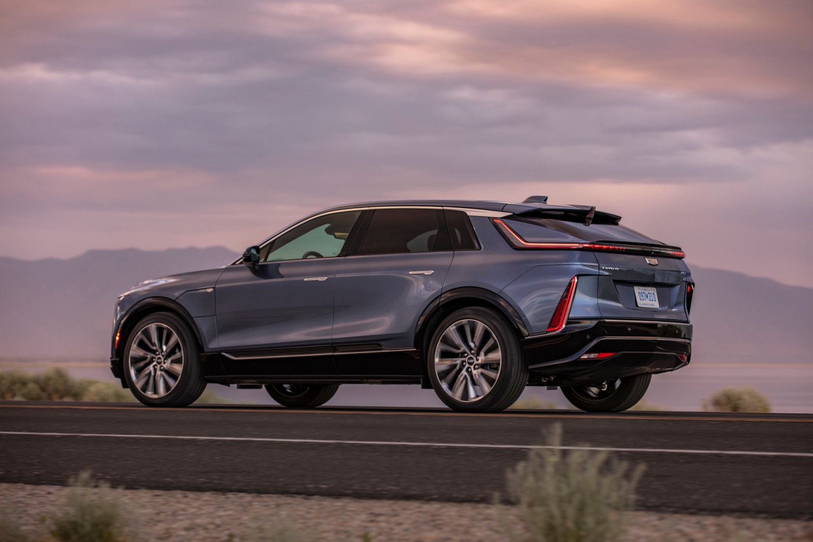 2026 Cadillac Lyriq - Rear three-quarter view of a blue Cadillac Lyriq driving on a road at sunset - Applewood Cadillac - New car dealership in Mississauga, Ontario