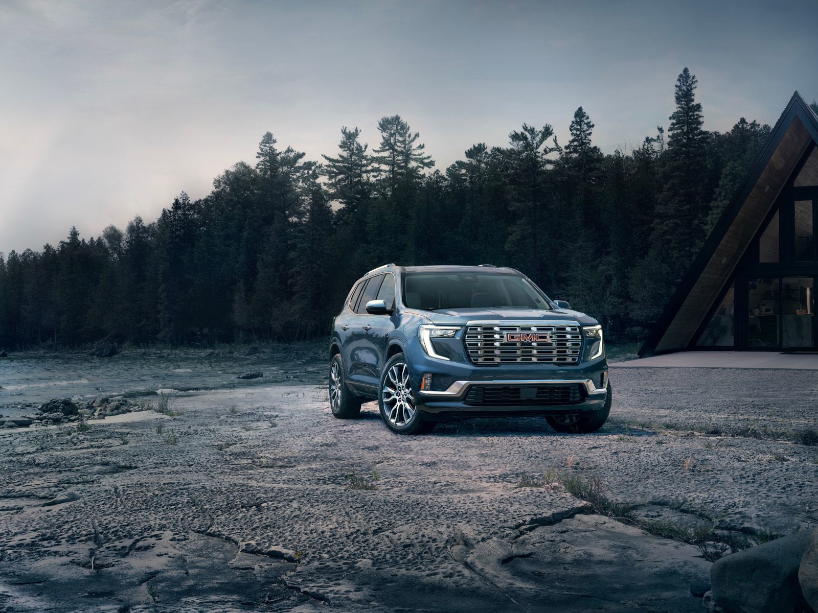 2026 GMC Acadia - A blue GMC Acadia parked on a rocky shoreline next to a modern A-frame cabin - Humberview Group - new car dealerships in Ontario