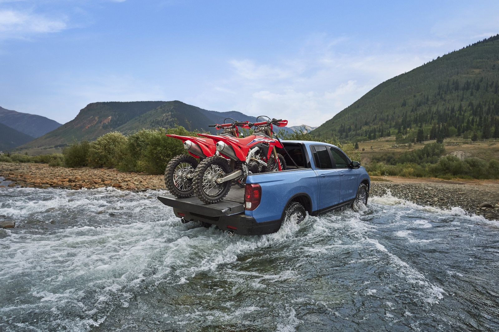2026 Honda Ridgeline - rear three-quarter view of a blue 2026 Honda Ridgeline crossing a shallow river with two dirt bikes in the bed - Orangeville Honda - New car dealership in Orangeville, Ontario