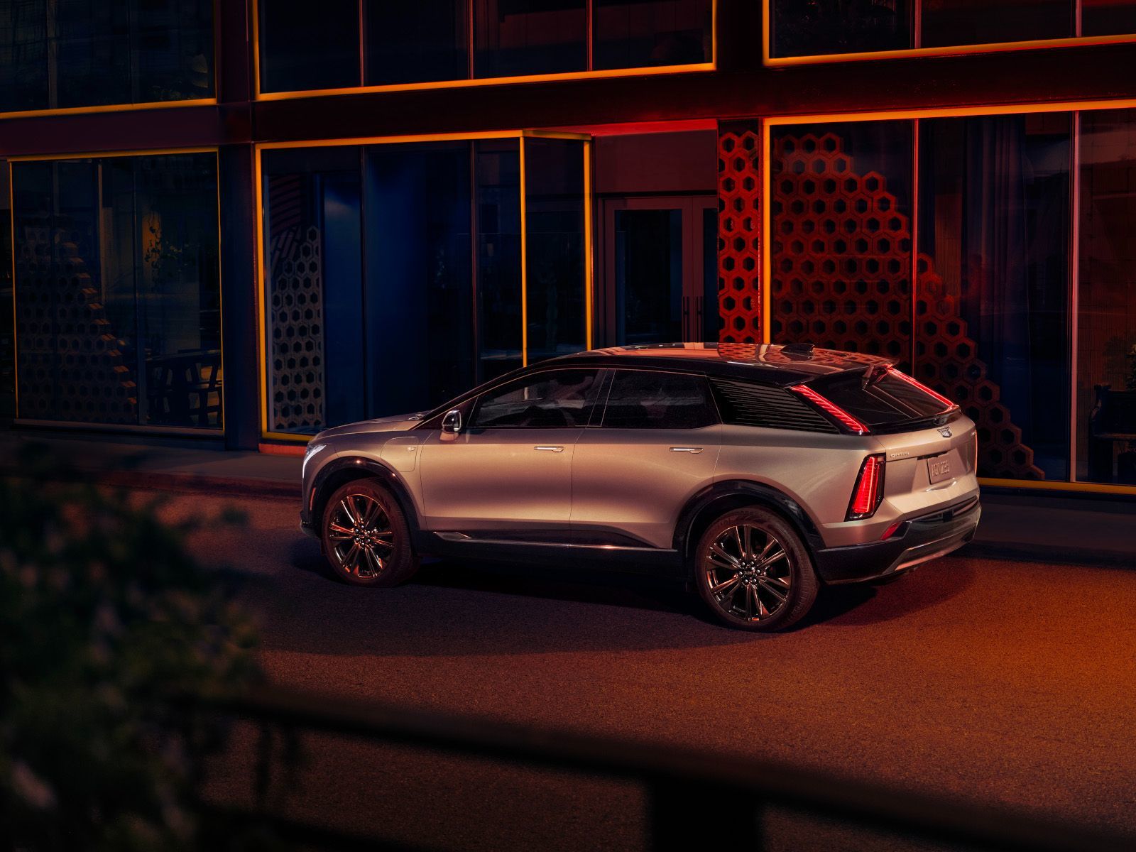 2026 Cadillac OPTIQ - rear three-quarter view of a silver Cadillac OPTIQ parked on a city street at night in front of a modern building with geometric light patterns - City Cadillac - Cadillac new car dealership in Toronto, ON