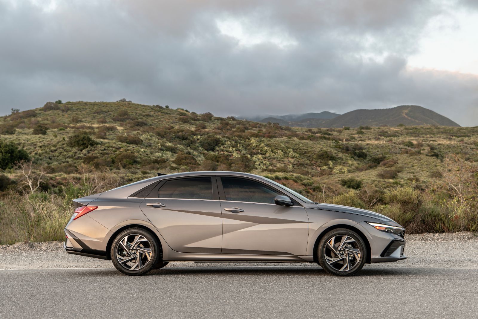 2026 Hyundai Elantra - A side profile view of a grey 2026 Hyundai Elantra sedan driving on a road with hills in the background - Woodstock Hyundai - New car dealership in Woodstock, Ontario