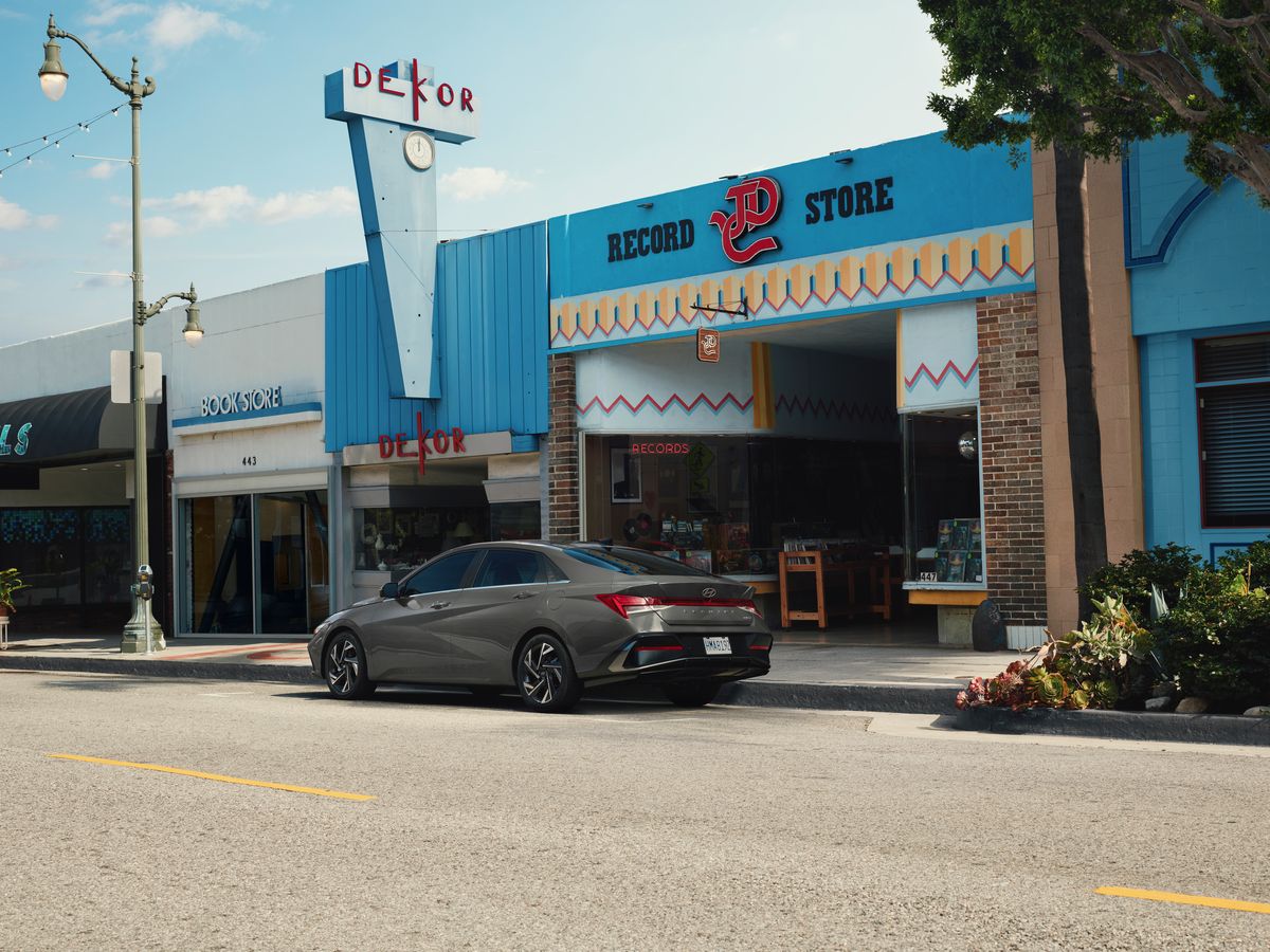 2026 Hyundai Elantra - A rear three-quarter view of a grey 2026 Hyundai Elantra sedan parked on a street near vintage storefronts - Woodstock Hyundai - New car dealership in Woodstock, Ontario