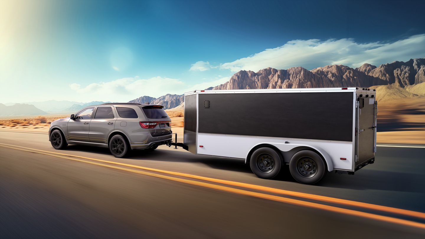 2026 Dodge Durango - A silver Dodge Durango towing a large enclosed trailer on a highway through a desert landscape - Cooksville Chrysler, Jeep, Dodge - New car dealership in Mississauga, Ontario