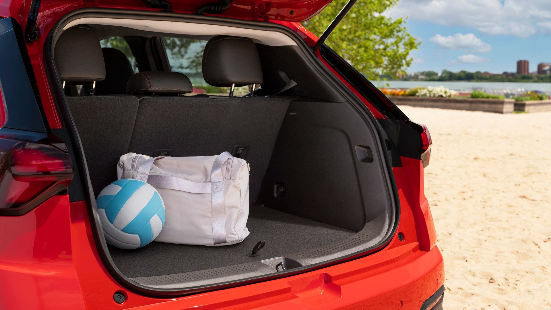 2027 Chevrolet Bolt - Close-up view of the open trunk of a red Chevrolet Bolt holding a duffel bag and a volleyball with a sandy beach in the background - Applewood Chevrolet, Buick, GMC - New car dealership in Mississauga, Ontario