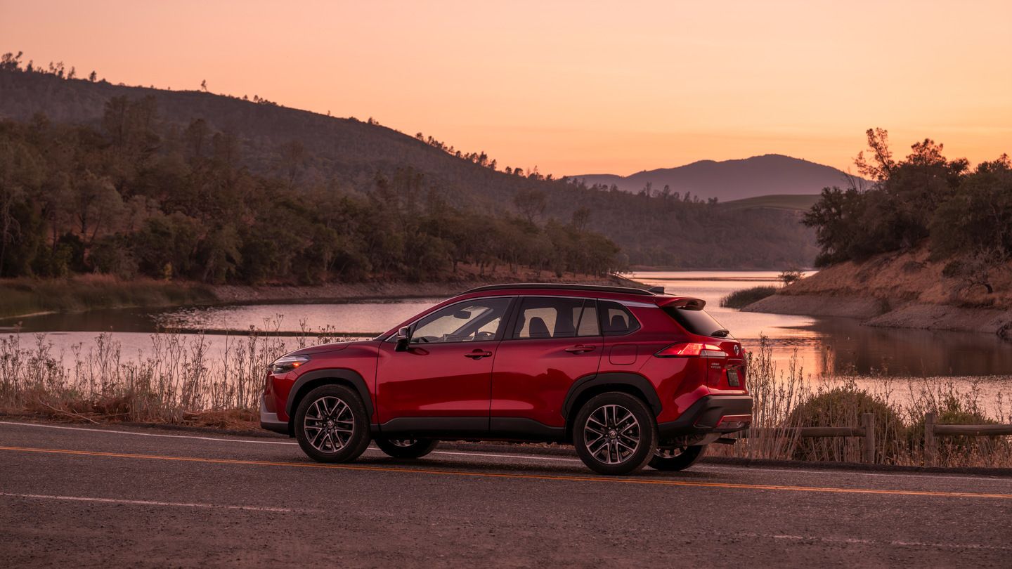 2026 Toyota Corolla Cross - A side view of a red 2026 Toyota Corolla Cross parked on a roadside with a lake and mountains in the background during sunset - Orangeville Toyota - Toyota car dealership in Orangeville, Ontario