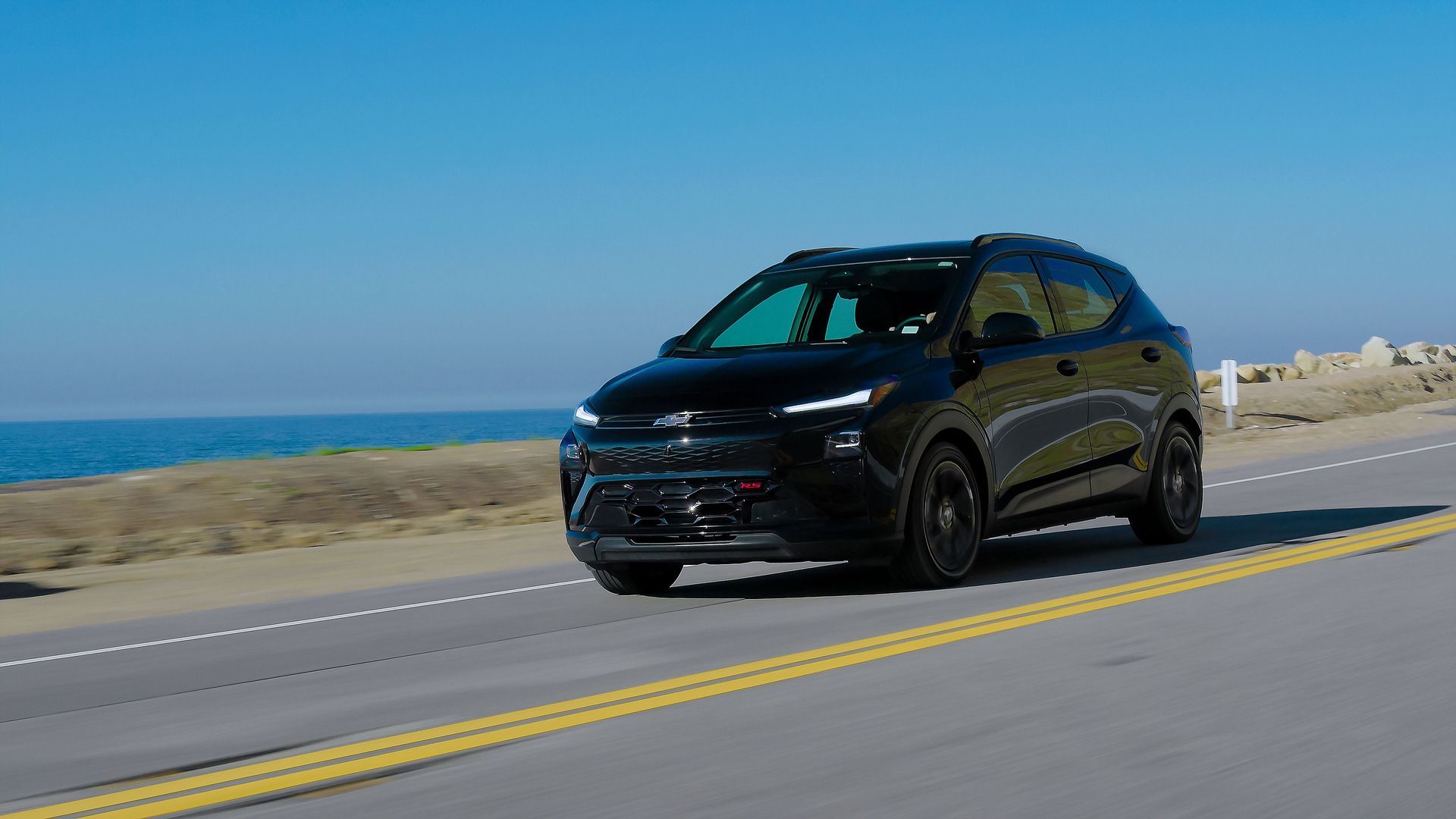 2027 Chevrolet Bolt - front three-quarter view of a black 2027 Chevrolet Bolt RS driving along an ocean road - Humberview Chevrolet, Buick, GMC - Chevrolet new car dealership in Toronto, Ontario