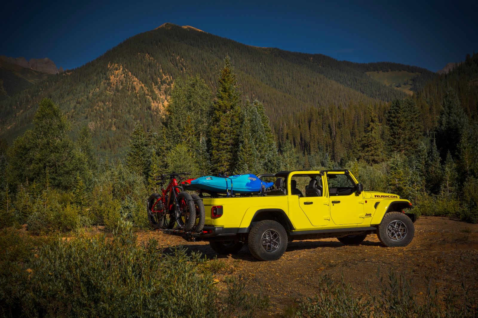 2027 Jeep Gladiator - rear side profile of a yellow 2027 Jeep Gladiator Rubicon loaded with mountain bikes and a kayak on an outdoor trail - Cooksville Chrysler, Jeep, Dodge, Ram - Jeep car dealership in Mississauga, Ontario