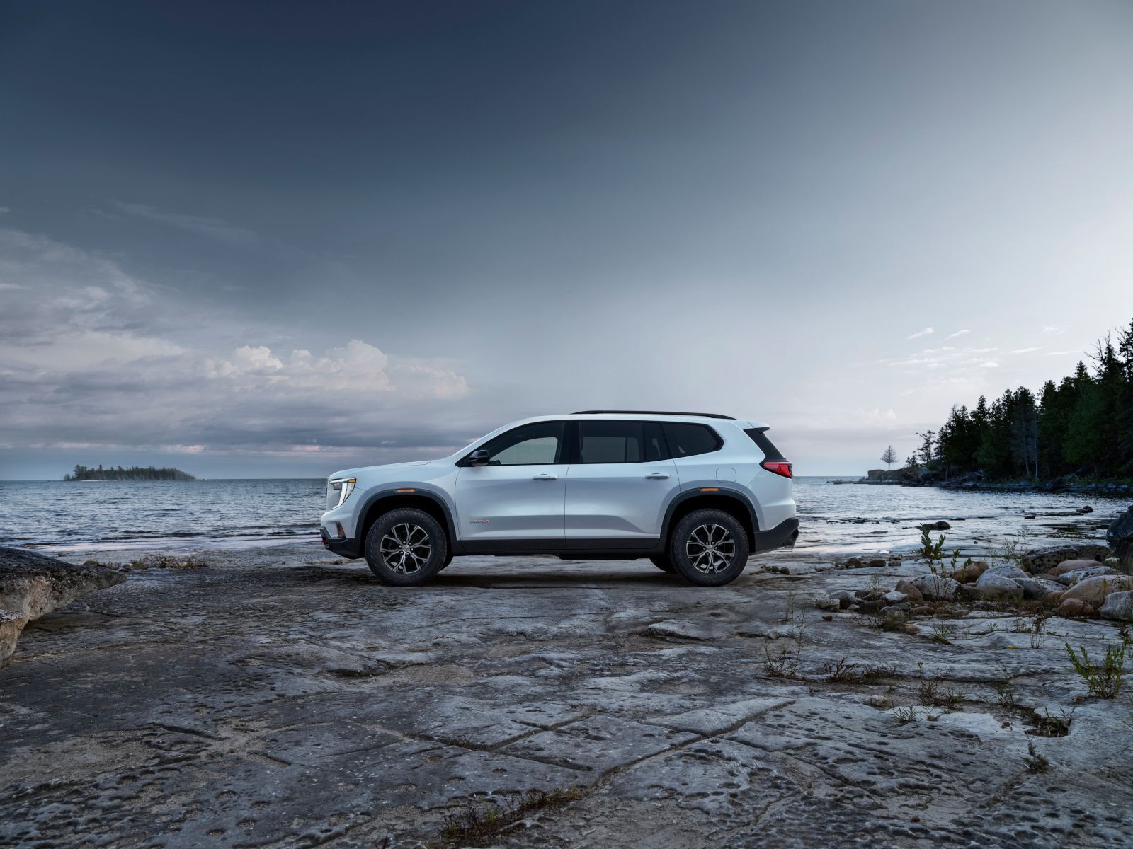 2027 GMC Acadia - side profile view of a white 2027 GMC Acadia AT4 parked on a rocky shoreline by the water - Applewood Chevrolet, Buick, GMC - GMC new car dealership in Mississauga, Ontario