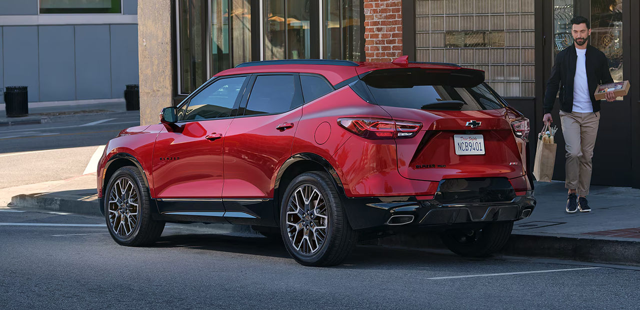 Vue 3/4 arrière et latérale du Chevrolet Blazer 2025, garé en bor de rue avec un homme se dirigeant vers la valise avec son épicerie
