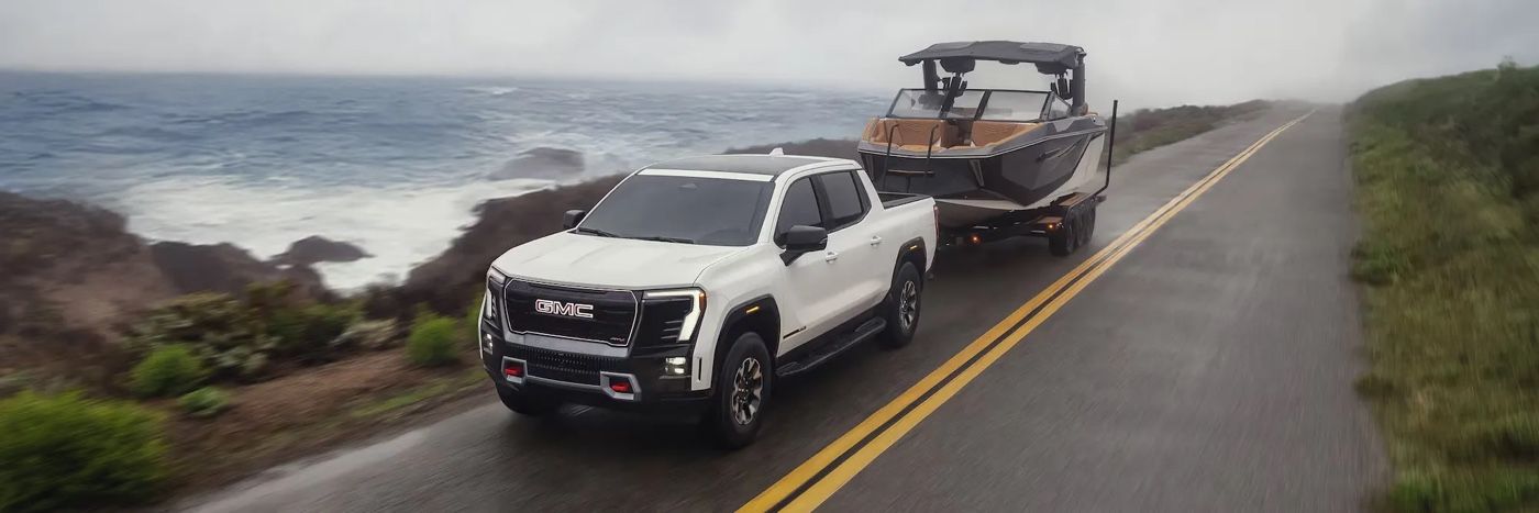 GMC Sierra EV 2026 blanc remorquant un bateau sur une route côtière brumeuse.
