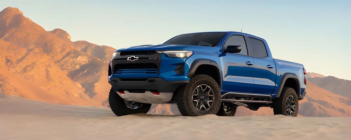 Pickup Chevrolet Colorado 2026 bleu stationné sur le sable devant des montagnes au lever du soleil.
