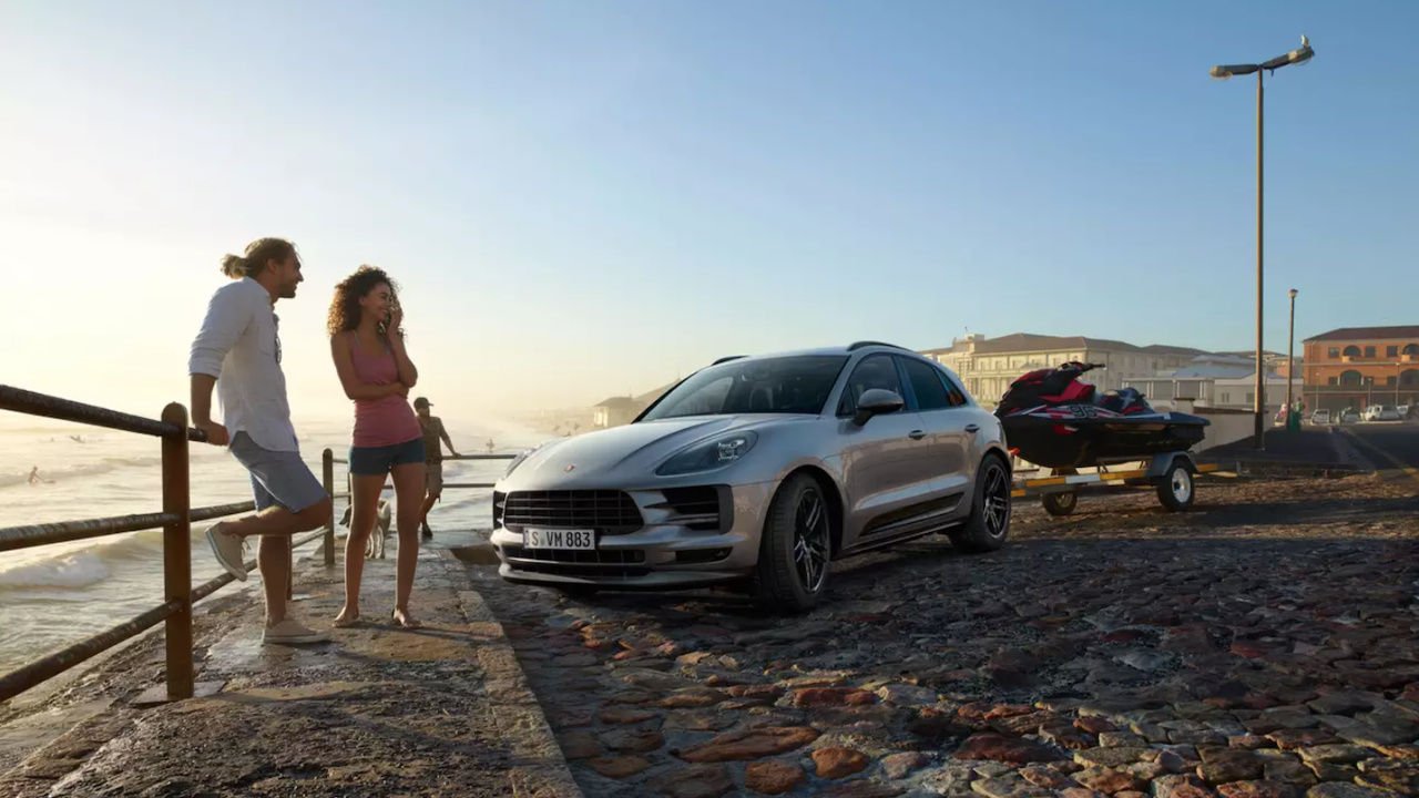 young couple by the sea in front of their 2023 Porsche Macan attached to a trailer carrying a jet ski