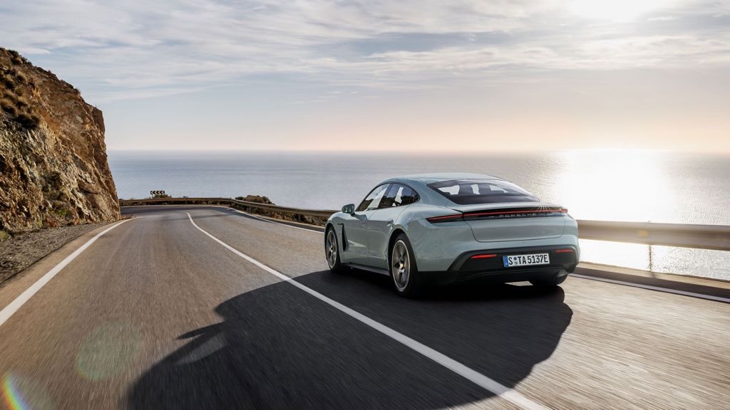 Rear 3/4 view of the 2025 Porsche Taycan electric car driving on a road by the sea.