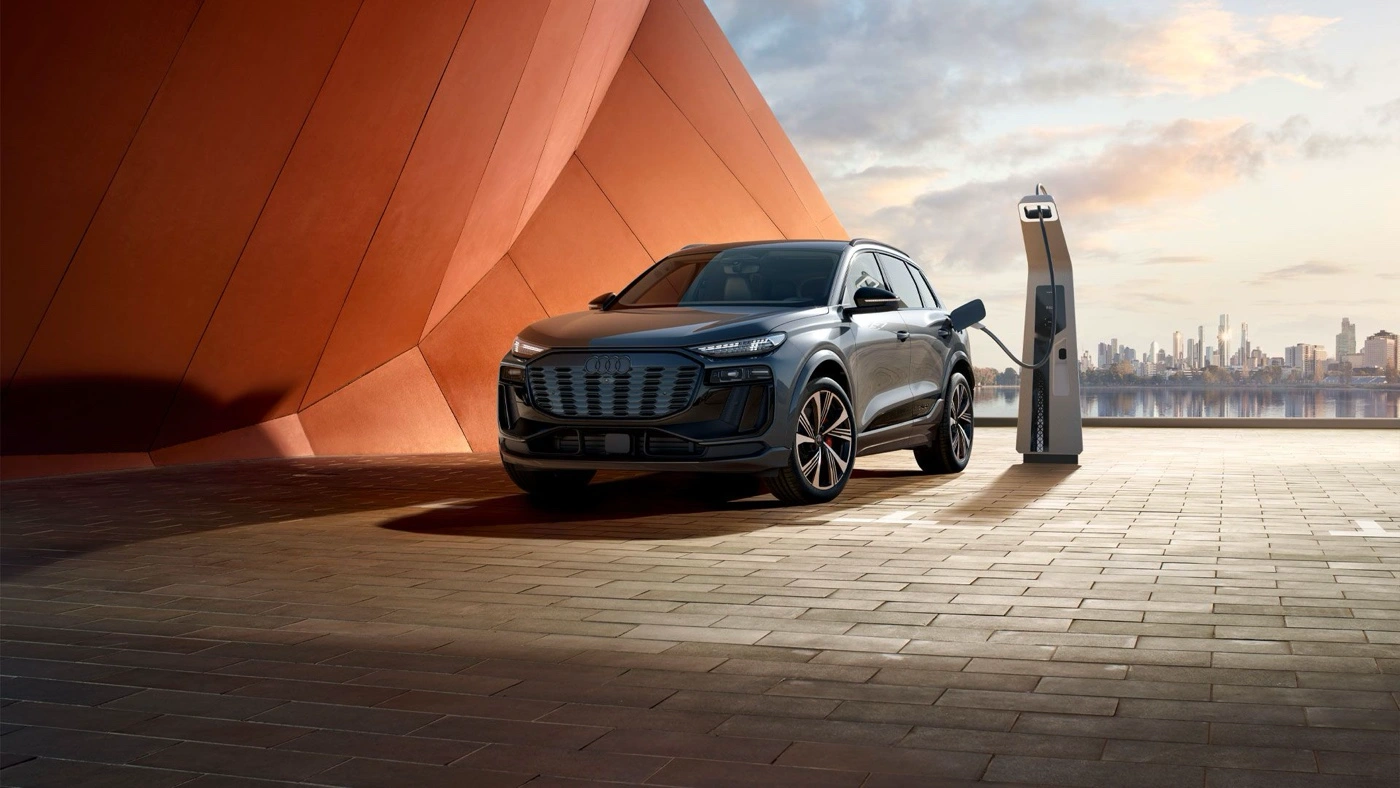 Version SQ6 de l&rsquo;Audi Q6 e-tron branch&eacute;e &agrave; une borne de recharge devant un d&eacute;cor urbain au coucher du soleil.