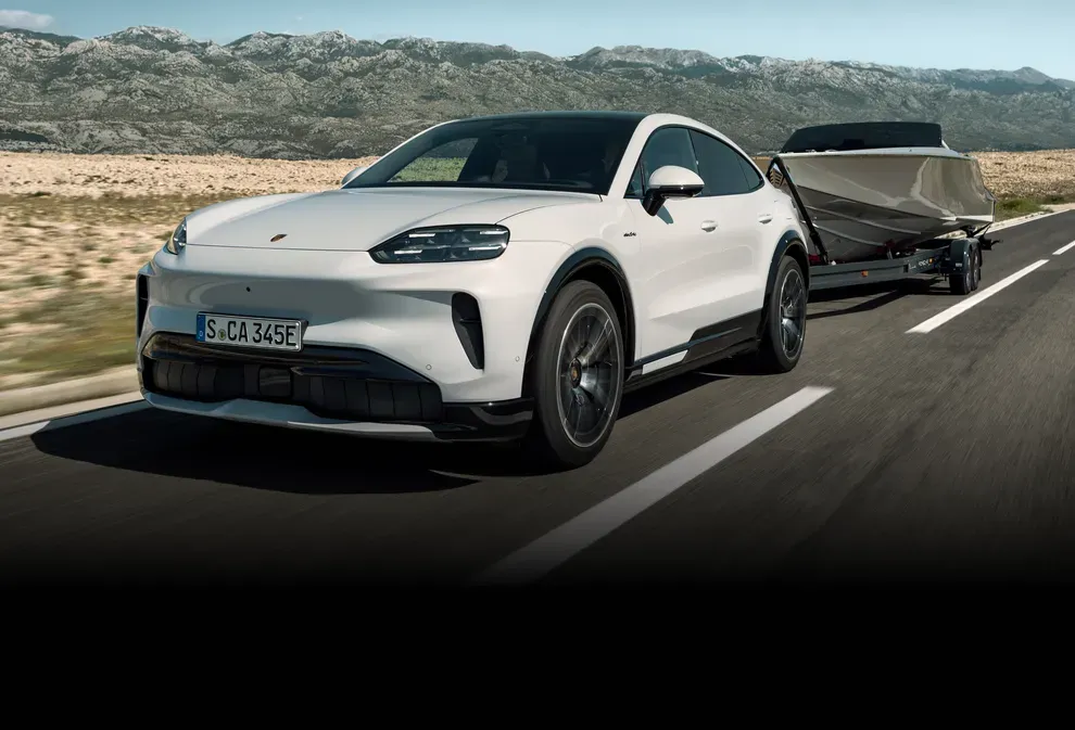 White 2027 Porsche Cayenne electric coupe towing a boat along a road.