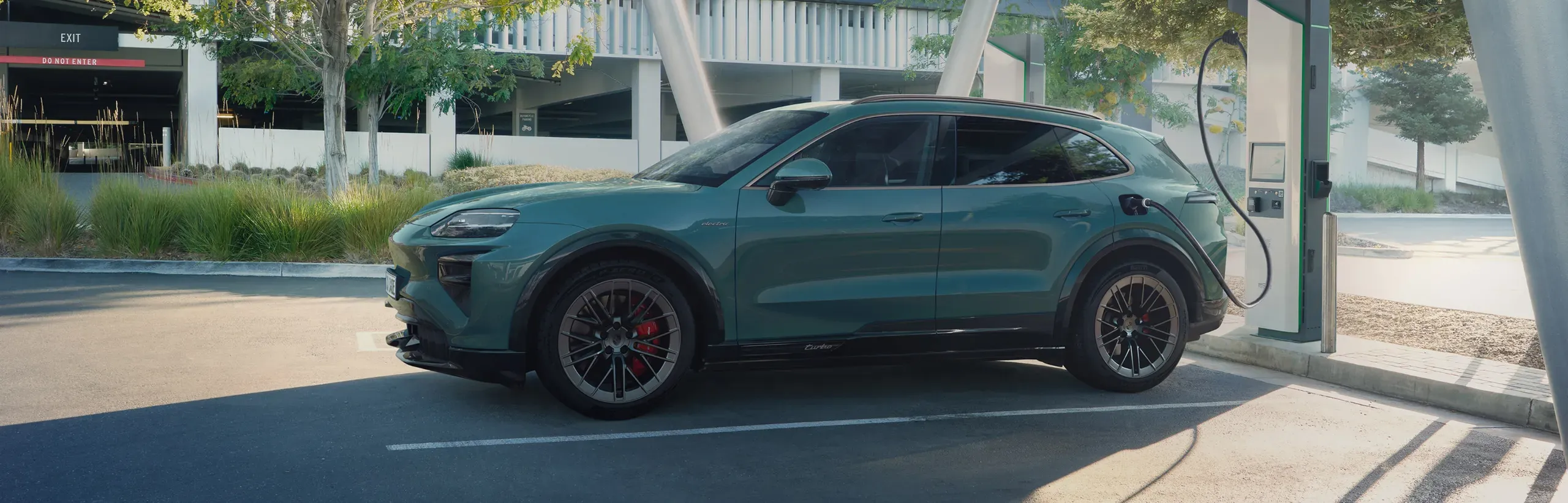 Blue-green 2027 Porsche Cayenne electric SUV shown in profile at a charging station.