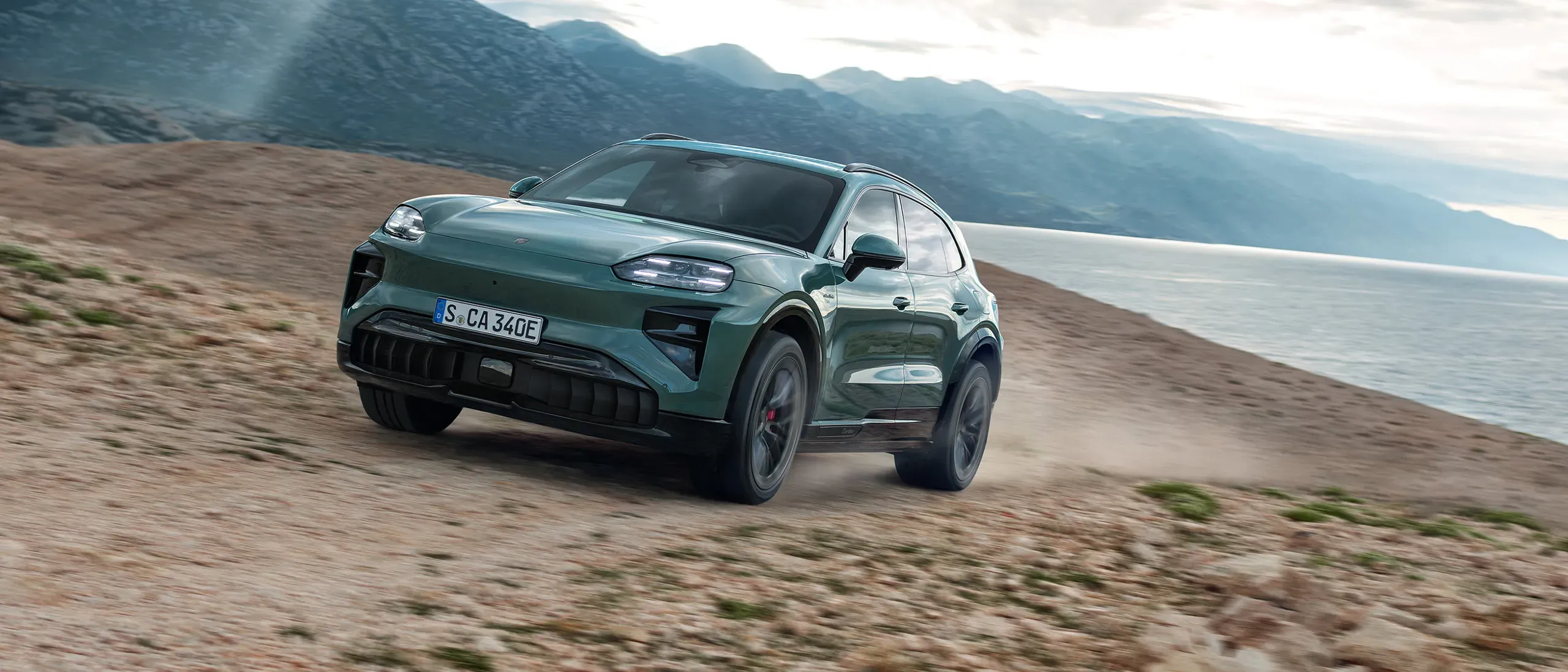 2027 Porsche Cayenne electric SUV on a dirt road beside a mountain lake.
