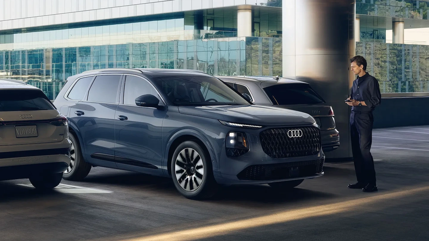 Blue 2027 Audi Q7 viewed from a front three-quarter angle, parked in front of a glass building.