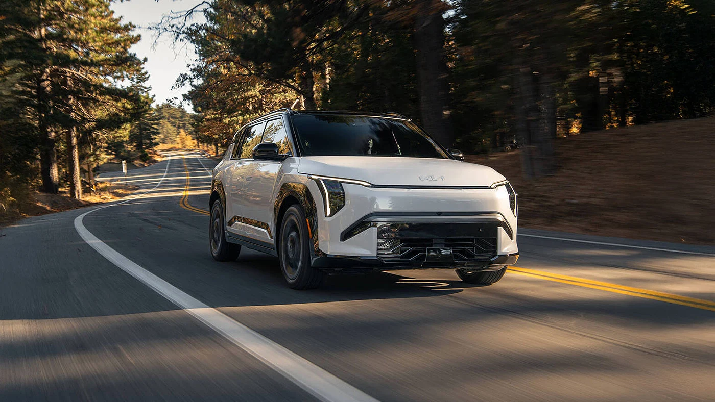 Kia EV3 2027 blanc en mouvement sur une route de campagne bord&eacute;e de conif&egrave;res