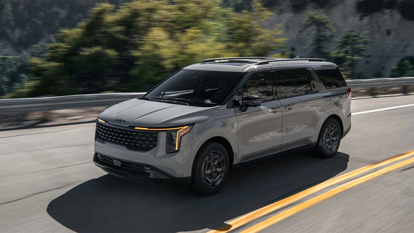 Kia Carnival 2026 gris en mouvement sur une route de montagne bord&eacute;e de conif&egrave;res