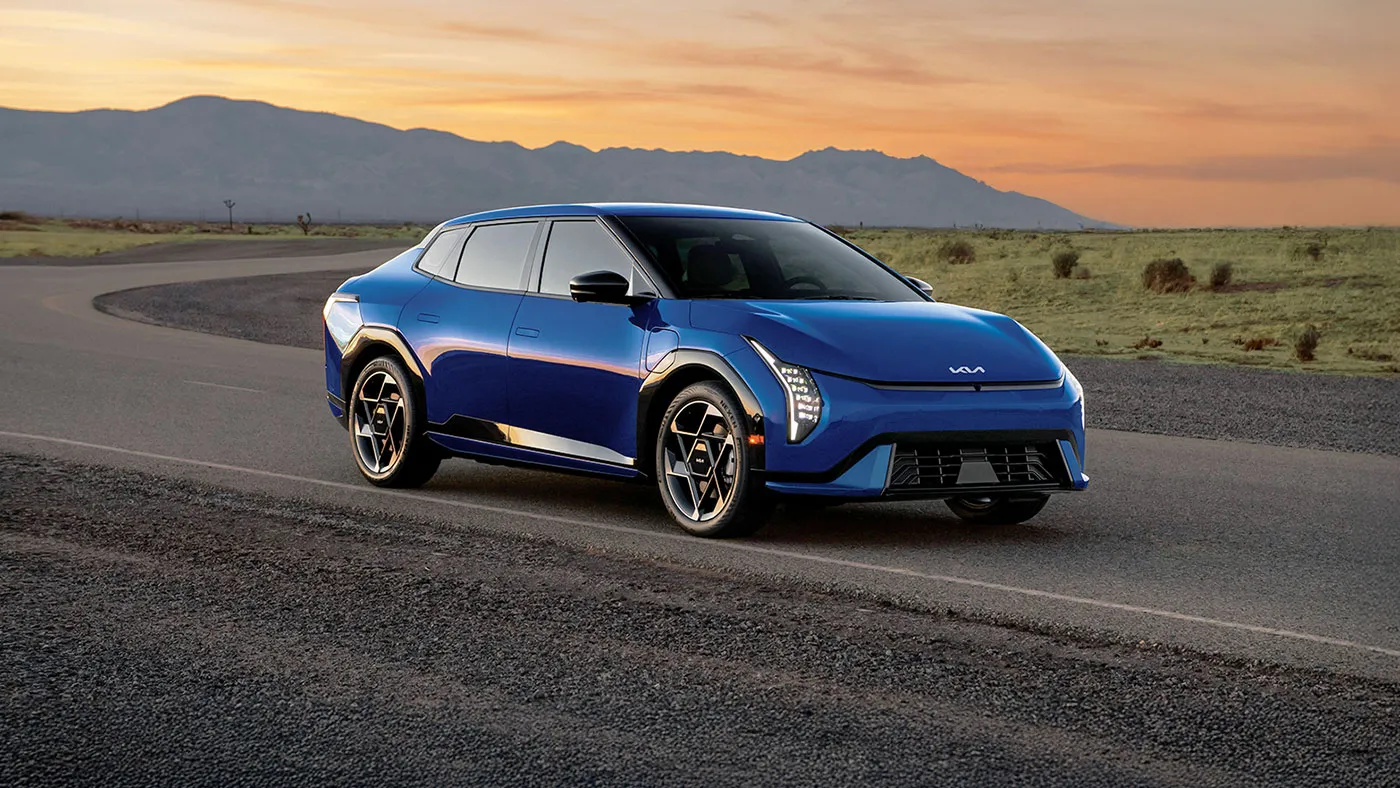 Kia EV4 &eacute;lectrique bleue vue de trois quarts avant sur une route au coucher du soleil.