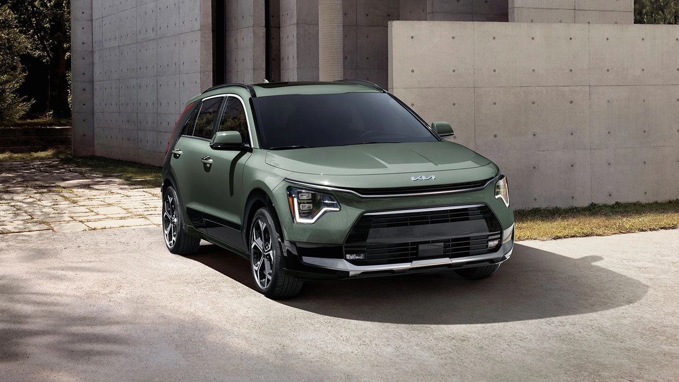 Vu 3/4 avant et lat&eacute;rale du Niro EV 2026 en arr&ecirc;t pr&egrave;s d'un bat&icirc;ment en b&eacute;ton