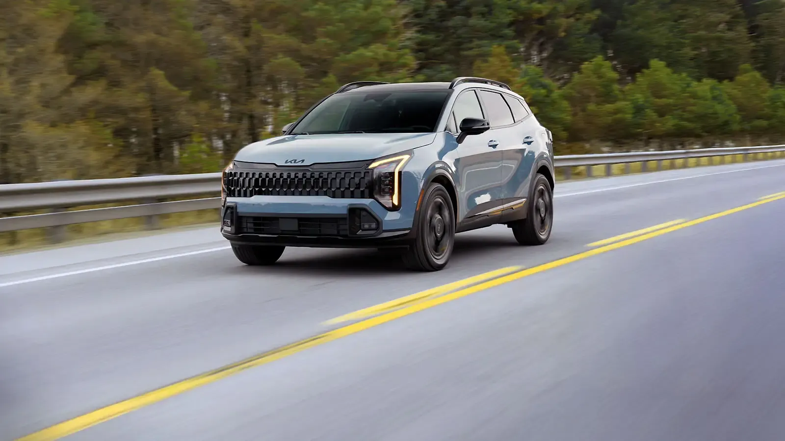 Kia Sportage 2026 bleu vu de trois quarts avant sur une route bord&eacute;e d&rsquo;arbres.