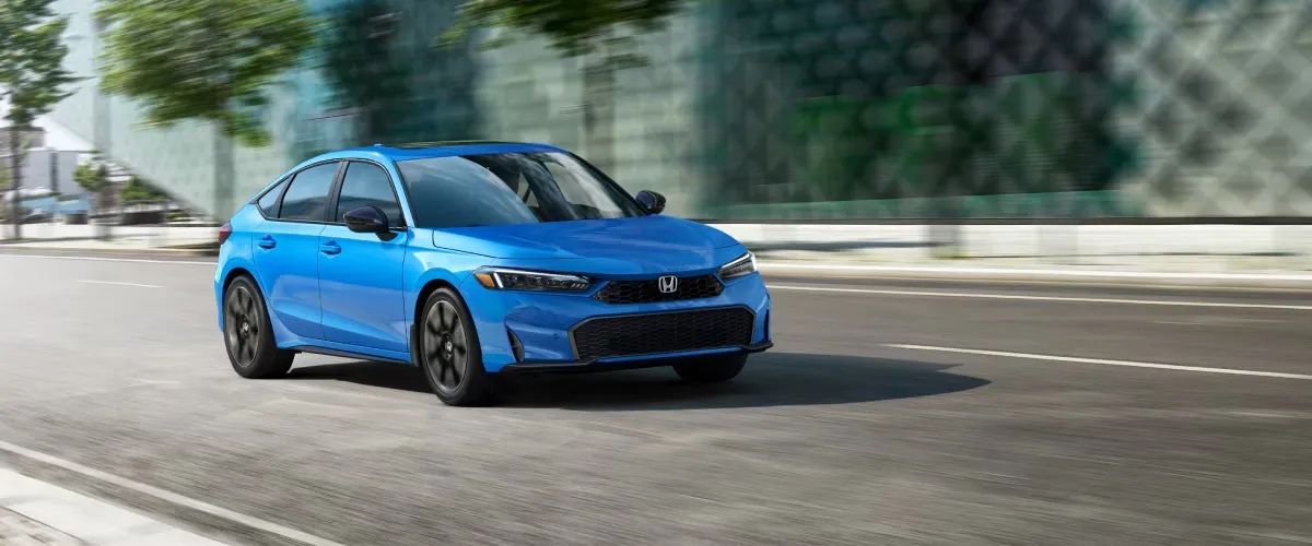 Honda Civic à hayon 2026 bleue en mouvement sur une route urbaine dégagée, avec fond flouté de bâtiments modernes et feuillage verdoyant.
