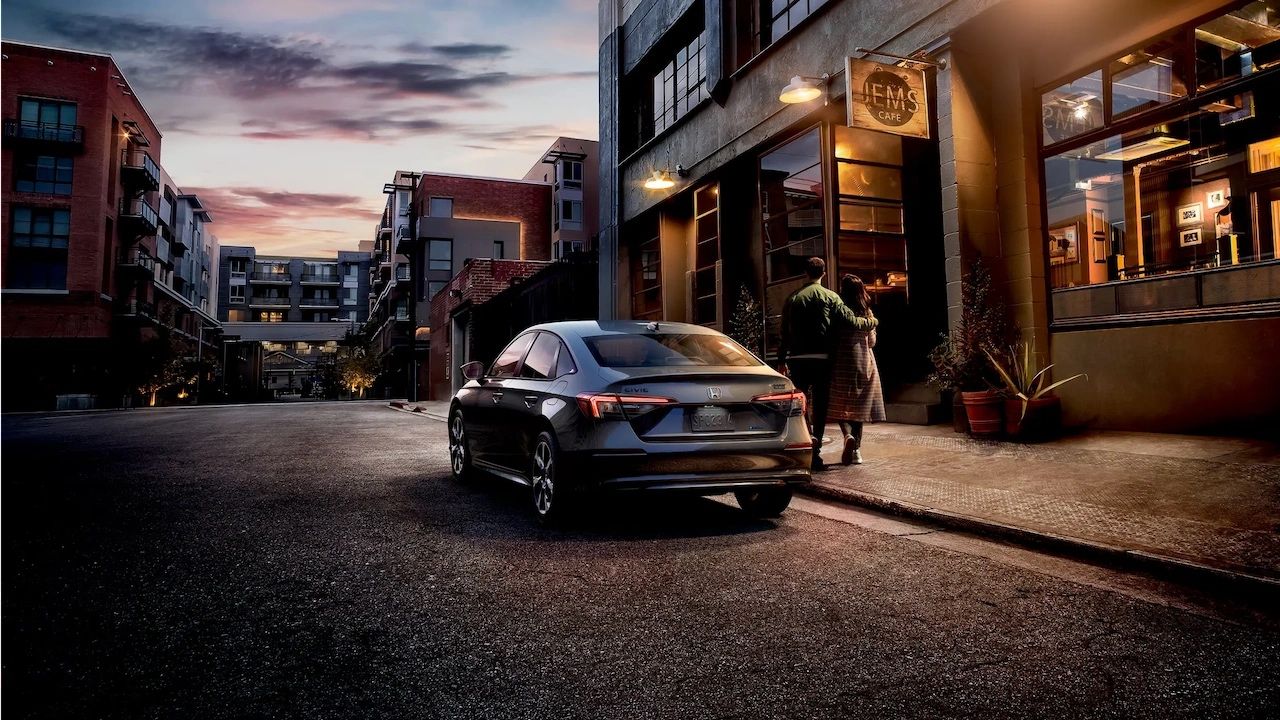 Vue arrière de la Honda Civic hybride 2026 stationnée devant un café urbain en soirée, avec un couple marchant sur le trottoir dans une ambiance chaleureuse et citadine.
