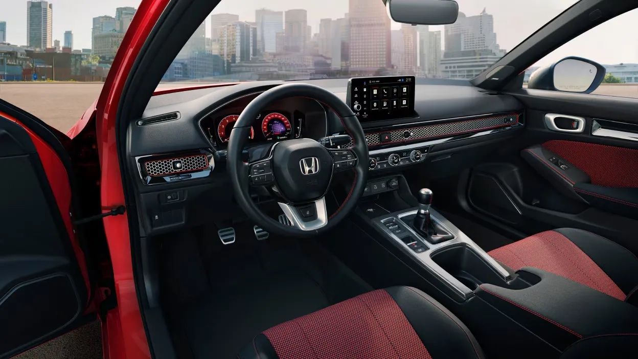 Habitacle de la Honda Civic SI 2026 rouge avec vue sur le tableau de bord sport, sièges avant en tissu rouge et noir, levier manuel et écran tactile central, avec arrière-plan urbain flouté.