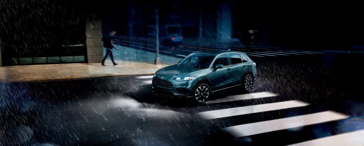 Honda HR-V 2026 vert foncé roulant sous la pluie de nuit, phares allumés et piéton traversant au loin.