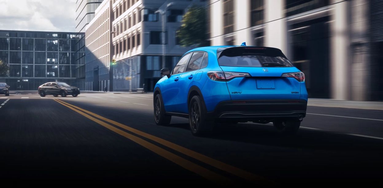Vue arrière du Honda HR-V 2026 bleu roulant sur une rue en ville, feux arrière visibles et design sculpté.