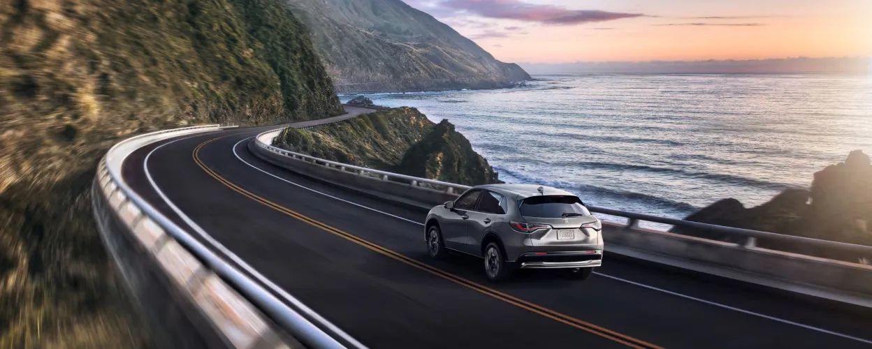 Honda HR-V 2026 gris roulant en bord de mer sur une route panoramique sinueuse au coucher du soleil.