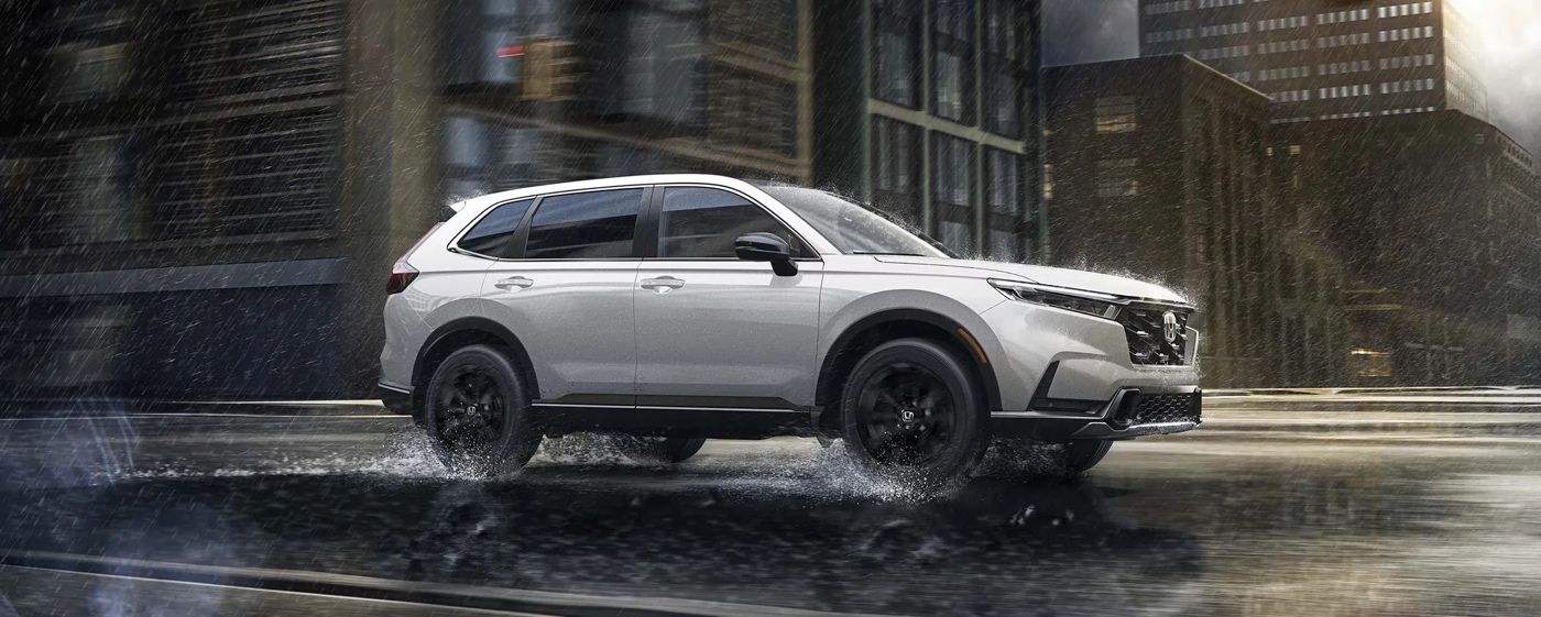 Vue latérale du Honda CR-V 2026 blanc roulant sous la pluie en ville, illustrant la traction et la stabilité du VUS compact sur chaussée mouillée.